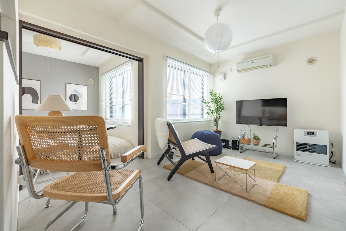 A well-lit living area features two stylish chairs and a small coffee table on a textured rug. A television is positioned against the wall, with a comfortable chair nearby. Large windows allow natural light to illuminate the space, complemented by neutral tones and decorative plants.