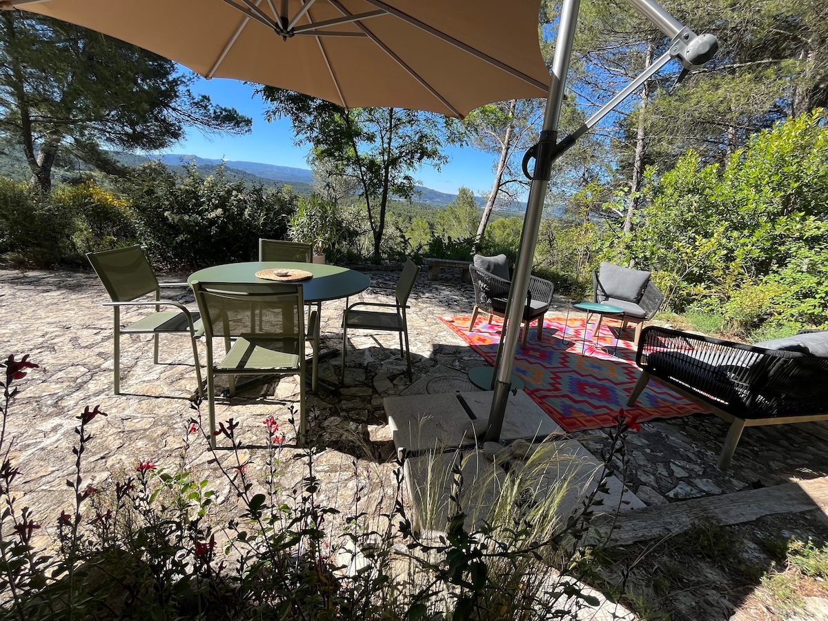 An outdoor terrace featuring a circular table surrounded by four chairs is highlighted. A large parasol provides shade, while a patterned rug lies beneath a seating area with comfortable chairs. Lush greenery and scenic views of the surrounding landscape can be seen in the background.