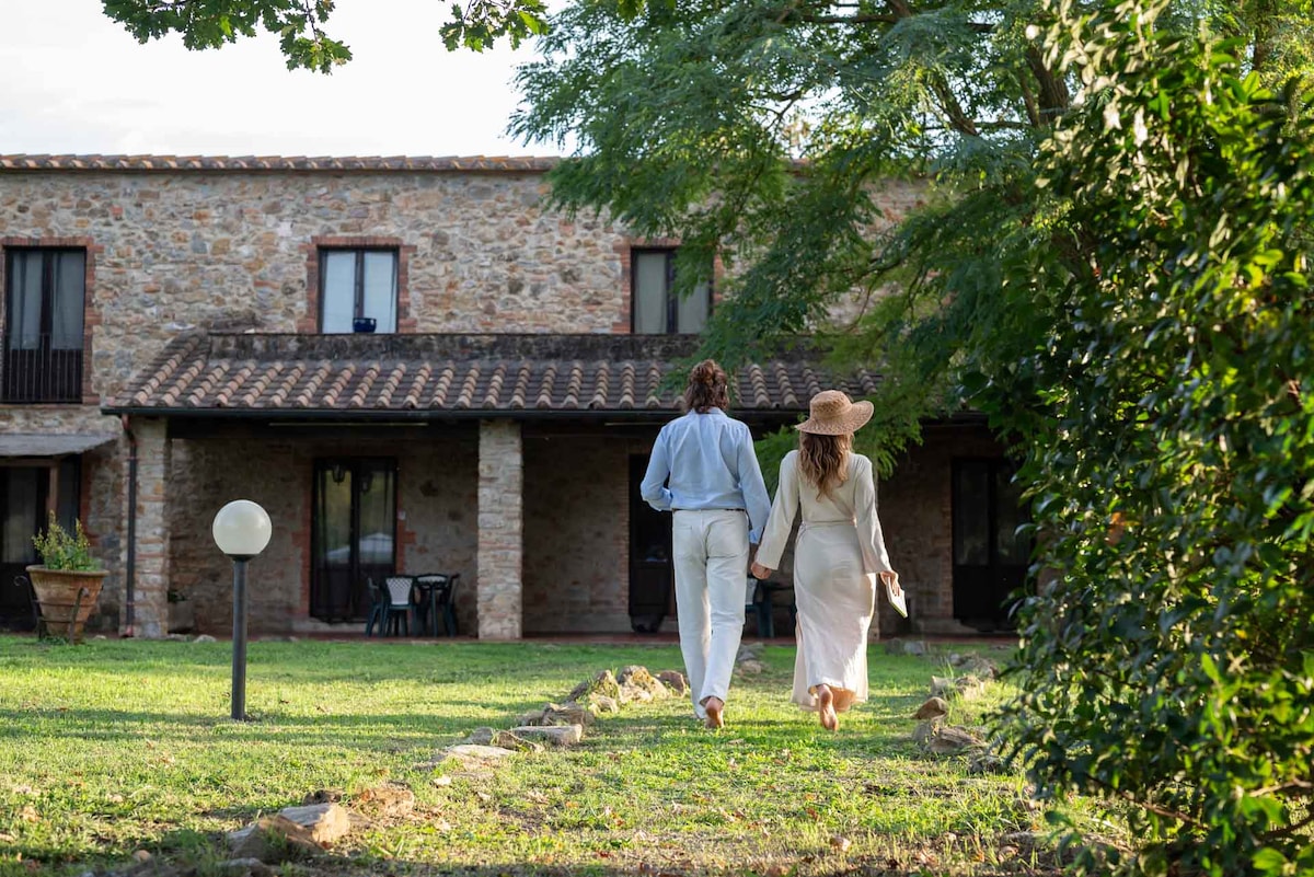 A couple is seen walking hand in hand along a path leading to a rustic stone building. The structure, featuring a tiled roof and multiple windows, is surrounded by well-maintained greenery. Outdoor seating is visible, creating a relaxed atmosphere.