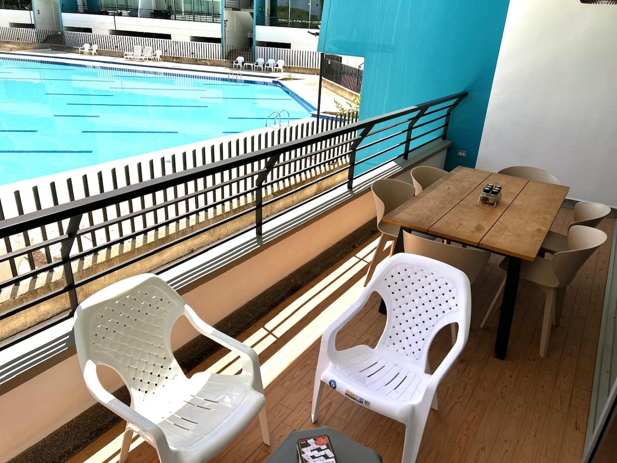 A balcony area is shown, featuring a table surrounded by four chairs. The space overlooks a communal swimming pool, with a barrier providing a clear view. The floor is wooden, and a small coffee table is positioned in front of the seating.