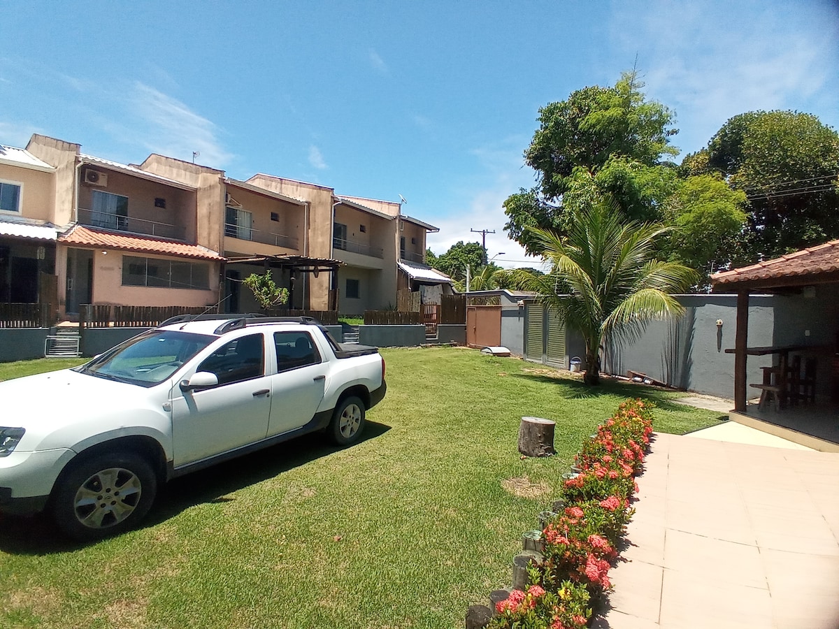A well-maintained outdoor area features a spacious lawn bordered by vibrant flowers. A white vehicle is parked on the grass, along with several residential buildings visible in the background under a clear blue sky. Surrounding vegetation includes a palm tree providing shade.