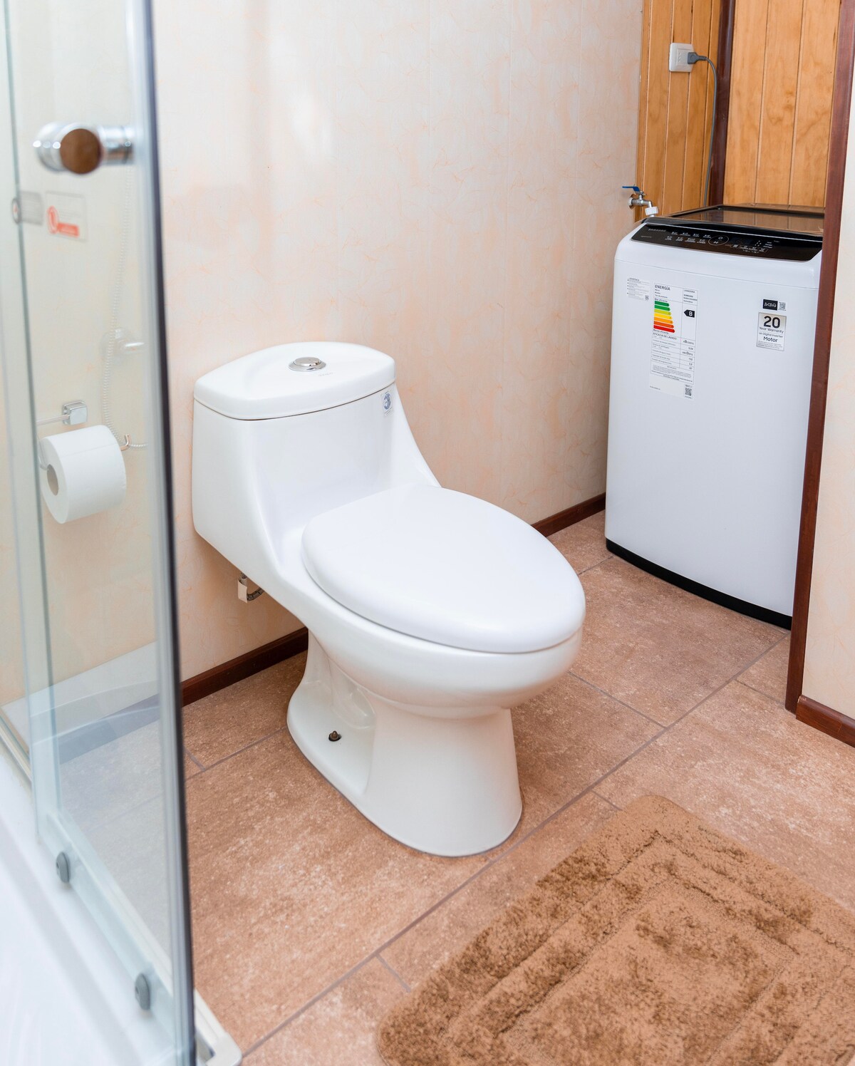 A clean bathroom is featured, showcasing a modern white toilet positioned next to a compact washing machine. Soft, neutral-colored tiles cover the floor, complemented by a light brown bath mat. A glass shower enclosure is visible, creating an airy and functional space.