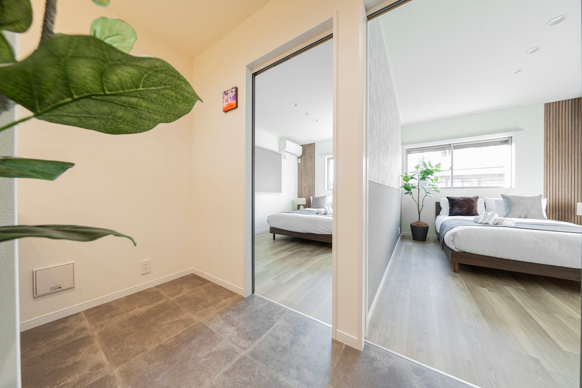 A spacious entrance area leads into a well-lit living space. A partially visible bedroom showcases a stylish bed with neutral bedding. Light-colored walls and wooden accents create a fresh and modern atmosphere, complemented by a green plant placed near the entrance.
