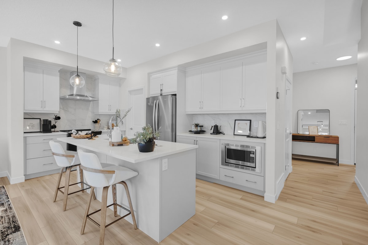 A modern kitchen features sleek white cabinetry and stainless steel appliances, including a refrigerator and microwave. A spacious island provides seating with three light-colored bar stools. Warm wood flooring complements the minimalist design, while natural light illuminates the open layout.