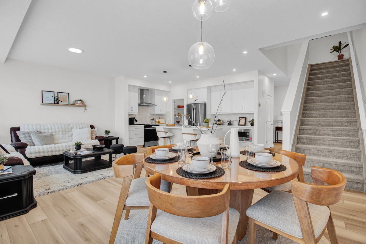 An open-concept dining area features a round table set for six with light-colored dishes. The modern kitchen is visible in the background, equipped with sleek cabinets and stainless steel appliances. A cozy seating area with a sectional sofa is positioned to the left, and stairs lead to the upper level.