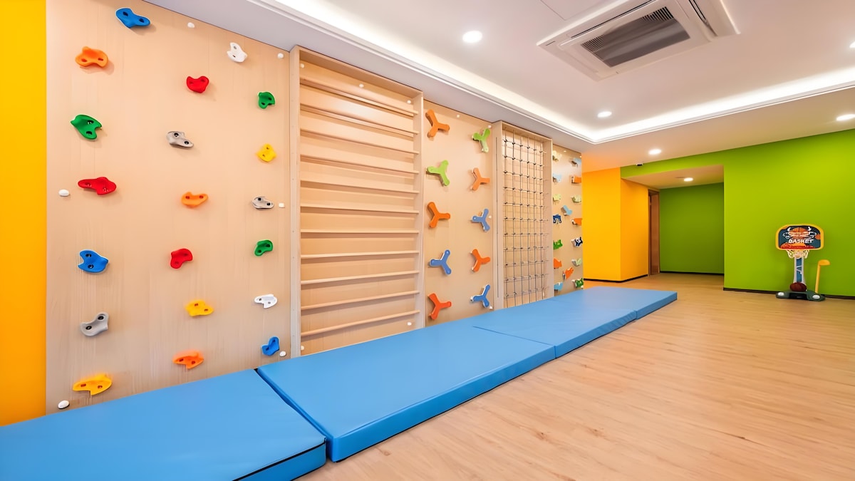 A colorful activity area features climbing walls with various grips in vibrant colors. Adjacent is a padded mat for safety. The walls are painted in bright hues, creating an engaging environment. Natural light floods the space through overhead lighting, enhancing the energetic atmosphere.