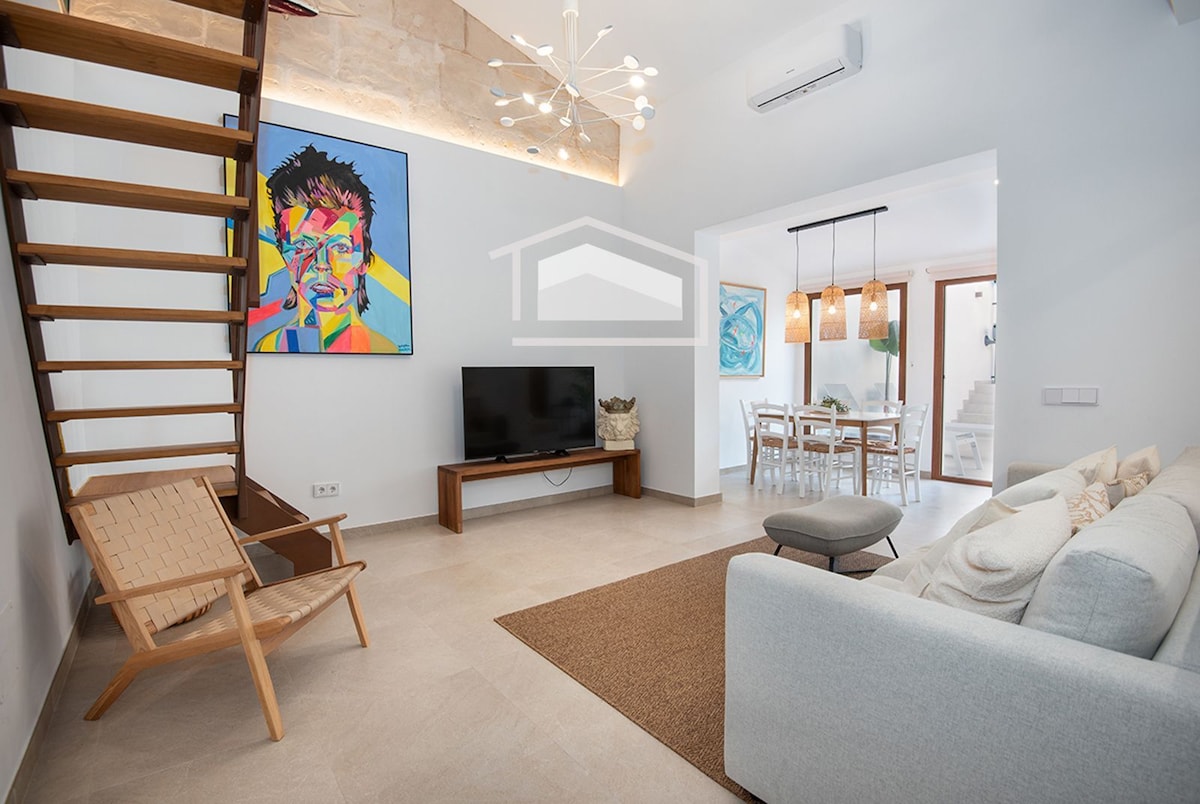 An open living area features a light-colored sofa and an armchair, complemented by a natural fiber rug. A modern artwork adorns the wall, while a flat-screen TV is positioned nearby. The space is illuminated by unique light fixtures, enhancing its airy feel.
