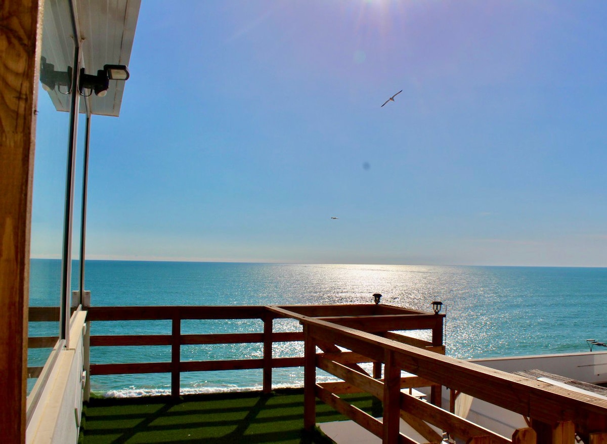 An expansive terrace provides unobstructed views of the shimmering ocean. The tranquil blue waters stretch to the horizon under a clear sky, with sunlight reflecting off the surface. A wooden railing encircles the area, complementing the outdoor space.