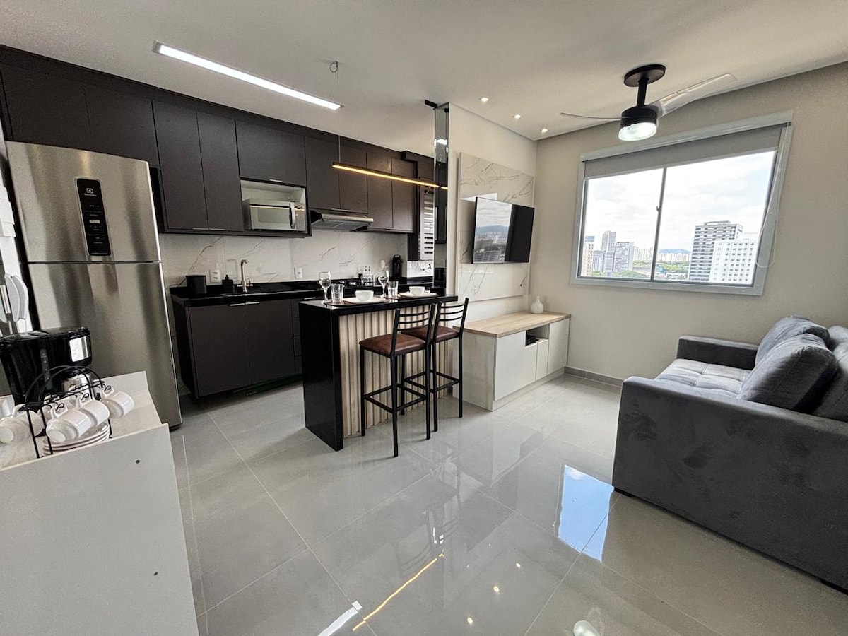 An open kitchen and living area features modern cabinetry and a countertop with seating for two. A comfortable sofa faces a wall-mounted television, and natural light enters through a large window. The sleek tiled flooring reflects the bright atmosphere.