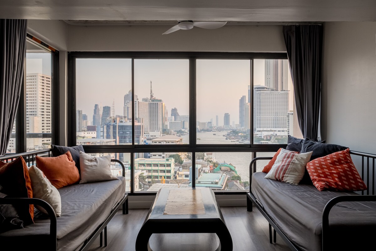 A spacious living area is presented with large windows offering expansive views of the river and city skyline. Two comfortable sofas are adorned with cushions in warm tones. A low coffee table occupies the center, enhancing the open layout filled with natural light.