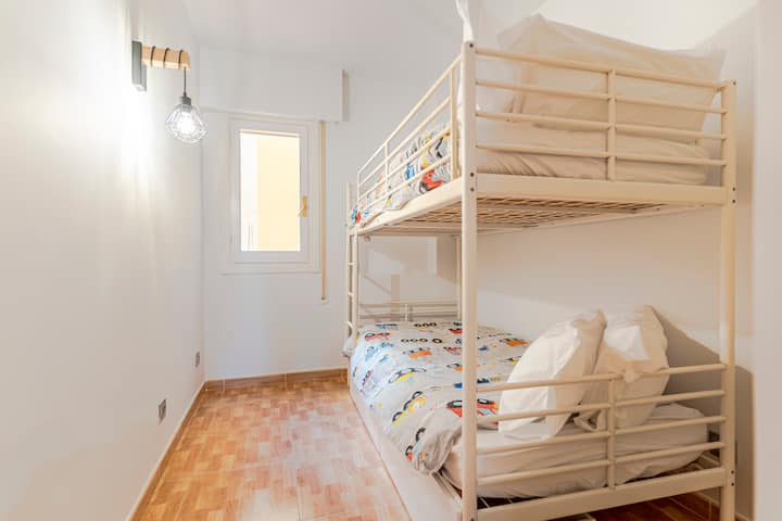 غرفة نوم مع سرير بطابقين معدني وسريرين مفردين، وفراش للأطفال، وخزانة مدمجة ونافذة مع ضوء طبيعي. أرضية باركيه وجدران بيضاء ومساحة عملية مثالية للأطفال أو الشباب.
