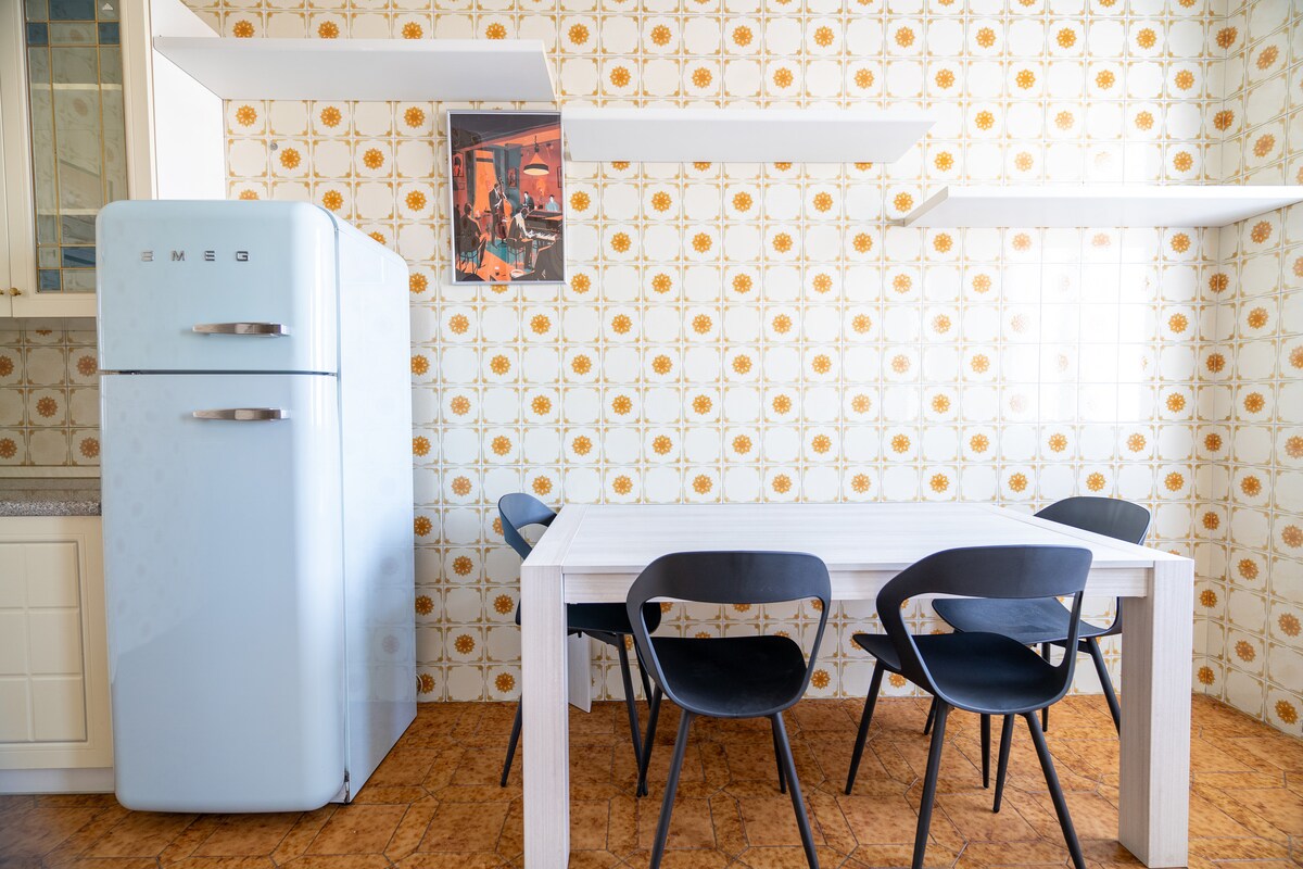 An inviting kitchen area features a retro-style refrigerator in pale blue and a white dining table surrounded by four black chairs. The walls are adorned with a patterned backsplash, complemented by open shelving above, creating a functional and aesthetically pleasing space.