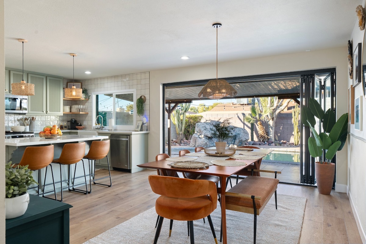 An inviting dining area features a large wooden table surrounded by comfortable leather chairs and a bench. Bright natural light flows in through accordion doors leading to an outdoor space, while modern pendant lights hang above the table, enhancing a functional yet stylish atmosphere.
