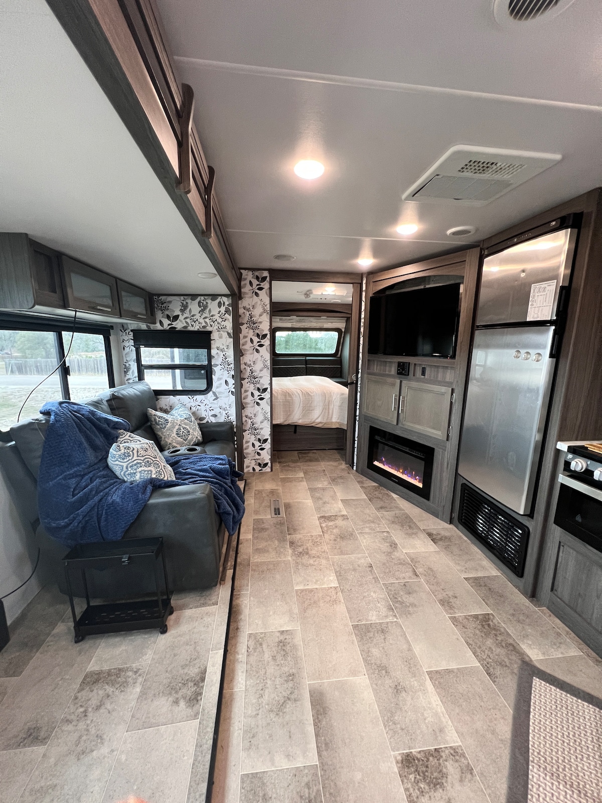 An inviting interior showcases a cozy seating area with a dark gray sofa adorned with patterned blue throw pillows. A compact fireplace adds warmth. The modern kitchen is equipped with stainless steel appliances, and the sleeping space is visible in the background through an elegant floral accent wall.
