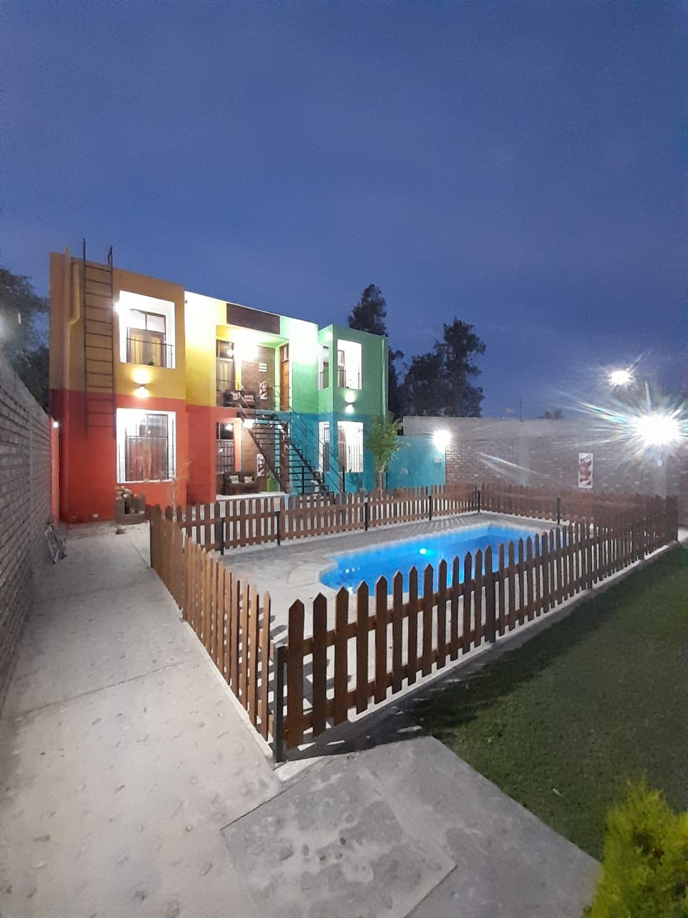 The exterior of the building features a colorful façade, outlined by a fenced yard containing a swimming pool. Dim outdoor lighting highlights the structure, while a paved walkway leads to the entrance. Lush greenery surrounds the area, providing a serene atmosphere.
