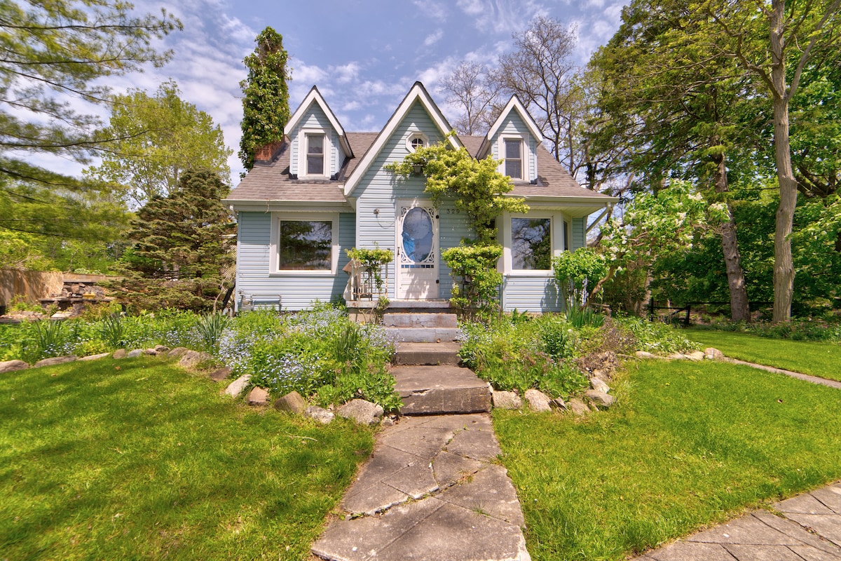 A charming house with a light blue exterior and a pointed roof is framed by lush greenery. A pathway of stepping stones leads to the front door, surrounded by colorful flowers and well-maintained grass. Mature trees provide shade, enhancing the serene outdoors.