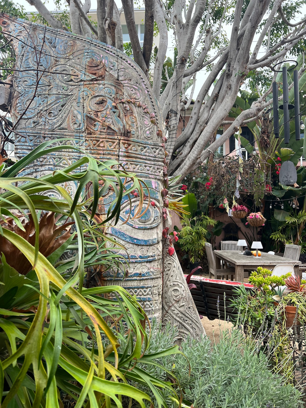 An intricately carved stone statue is surrounded by lush greenery, showcasing a variety of plants and flowers. A tranquil seating area is visible in the background, where wooden furniture complements the vibrant garden setting.