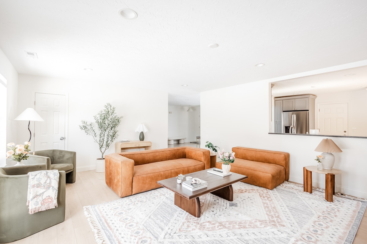 The living area features two stylish tan sofas arranged around a central coffee table, with a decorative area rug anchoring the space. A floor lamp and potted plant add a touch of greenery, while a pass-through to the kitchen can be seen in the background.