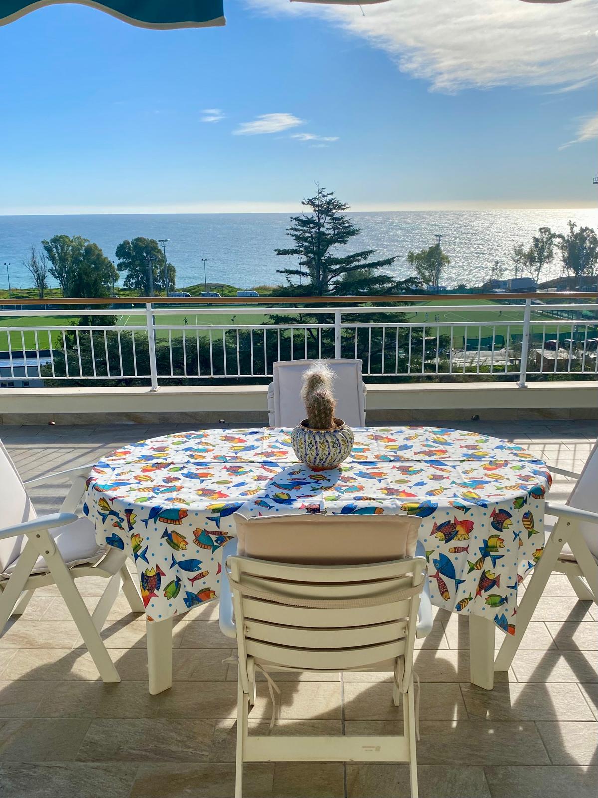 A round table is set on a balcony featuring a colorful tablecloth decorated with various fish. Four white chairs surround the table, while a decorative plant sits in the center. The sea is visible in the background, complemented by a clear sky and distant shoreline.