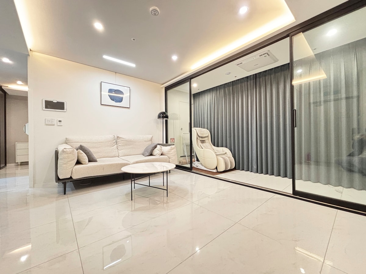 A spacious living area features a comfortable sofa and a round coffee table. Large glass doors lead to a room with floor-to-ceiling curtains, offering natural light. A recliner is positioned next to the curtains, enhancing the space's modern design.