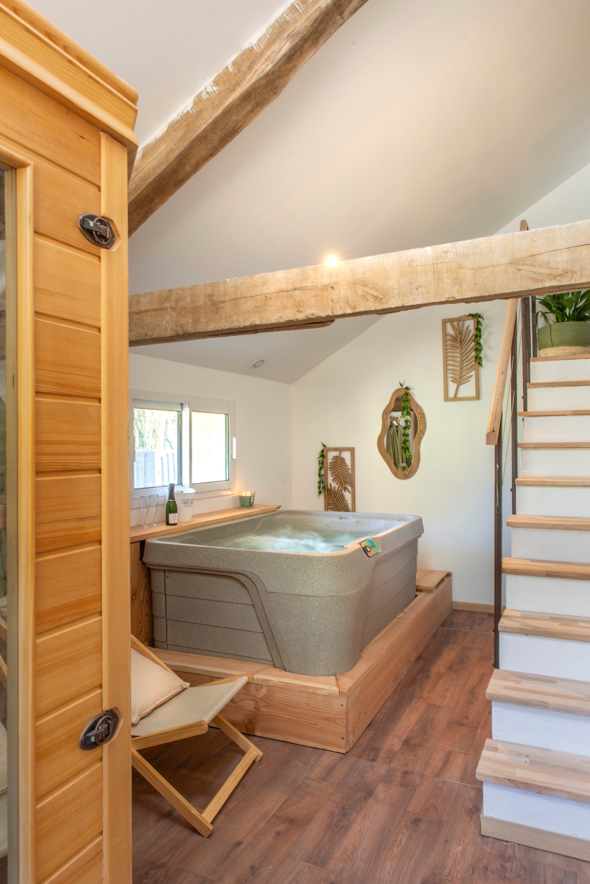 A serene relaxation area features a private Jacuzzi situated alongside a wooden sauna. Natural light enters through a window, illuminating the wooden beams above. A tastefully arranged plant adds a touch of greenery, and a comfortable chair is positioned nearby.