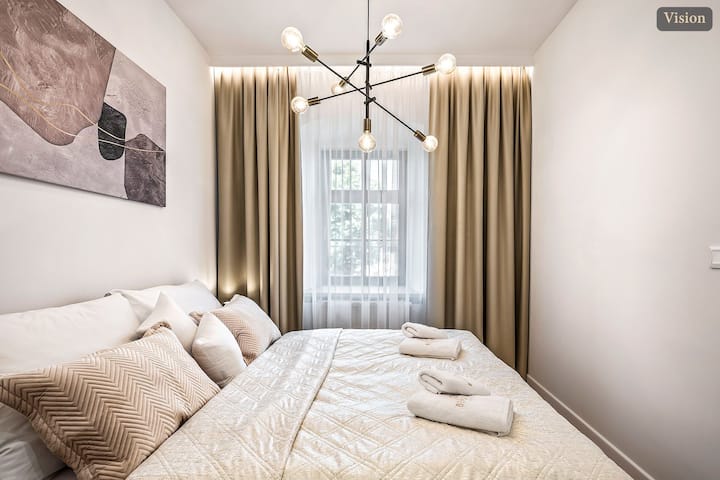 El dormitorio Vision muestra una cama tamaño king con almohadas en capas, ropa de cama suave en tonos neutros, toallas dobladas, un candelabro de diseñador, cortinas beige y una gran obra de arte sobre la cabecera. La ventana aporta una suave luz natural a la habitación.