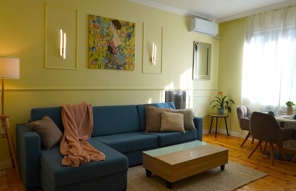 A living area features a light yellow wall with an abstract artwork displayed above a comfortable blue sectional sofa. A wooden coffee table sits centered in front of the sofa. Natural light streams through a large window, illuminating a small dining area set for four.