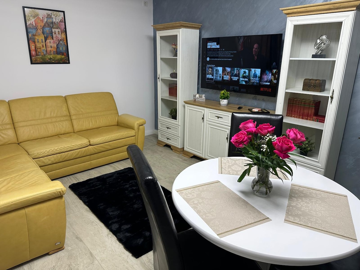 A modern living area features a yellow sectional sofa and a round dining table set with four black chairs. A vase of pink roses rests on the table. A television mounted on the wall displays content, while decorative cabinets flank either side, adding storage and aesthetic appeal.