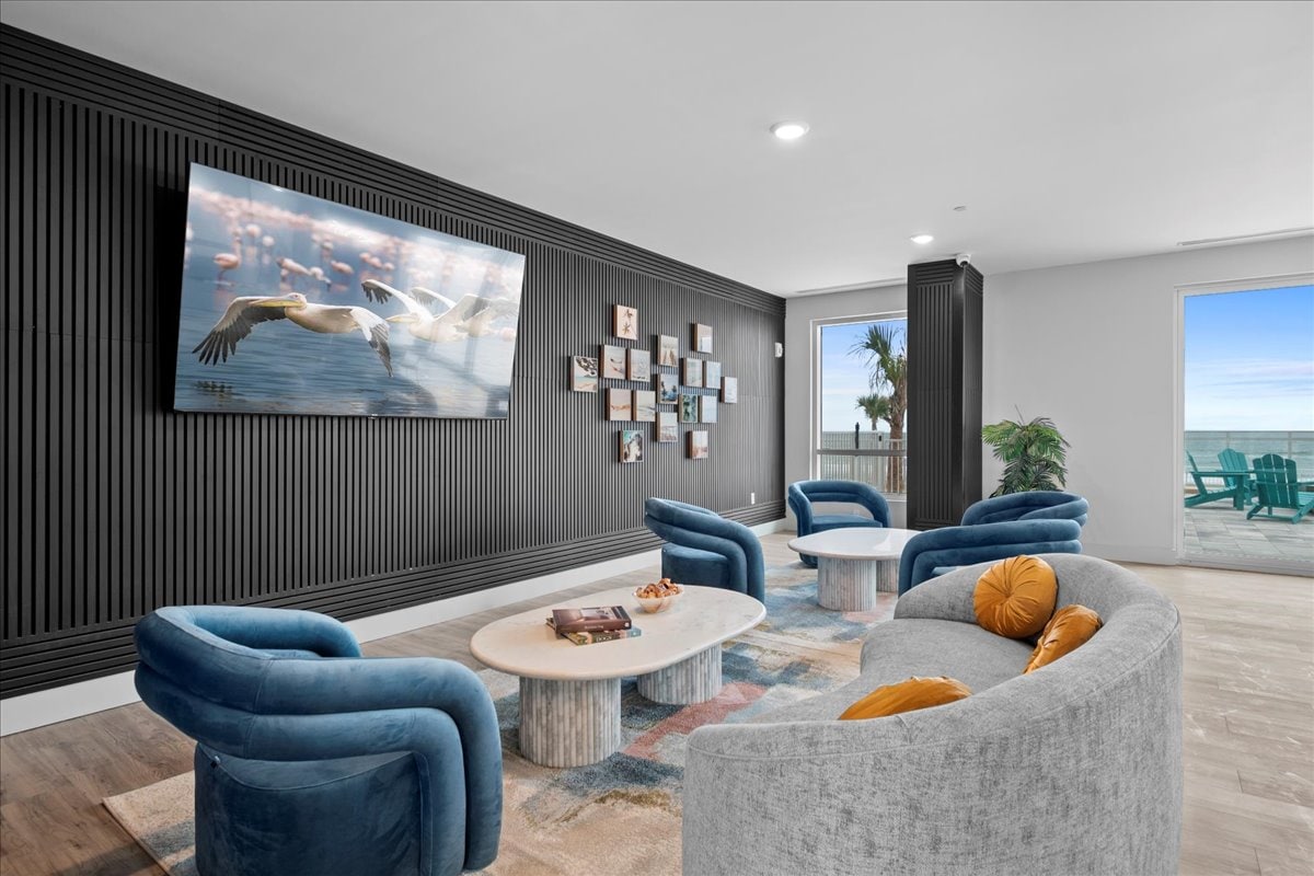 A modern lounge area features curved sofas and accent chairs, arranged around a circular coffee table. A large television is mounted on a textured wall adorned with framed photographs. Natural light floods in from floor-to-ceiling windows, revealing glimpses of the ocean beyond.