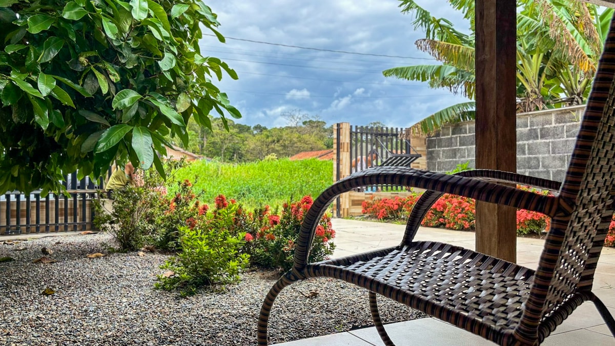 A shaded outdoor space features a comfortable wicker chair positioned on a stone patio. Lush green vegetation and colorful flowering plants surround the area, while a wooden fence and scenic landscape are visible in the background beneath a partially cloudy sky.