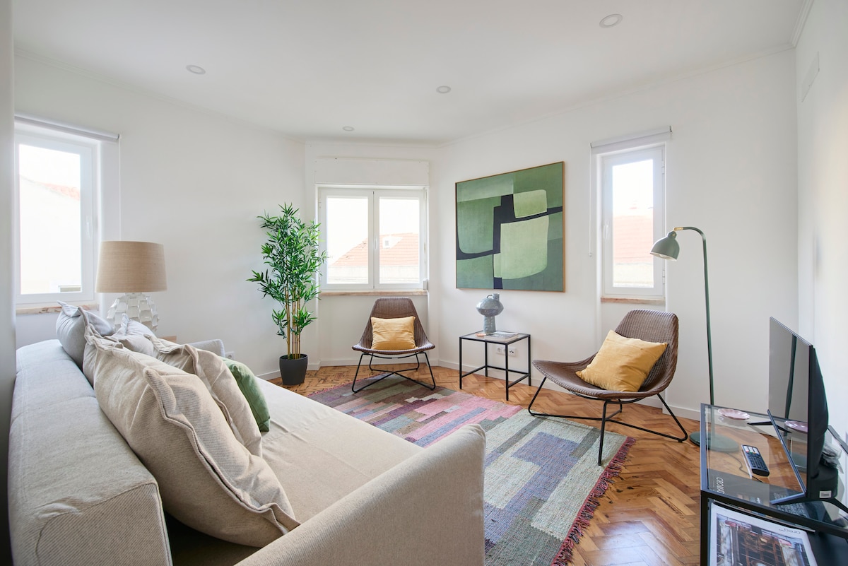 A bright living room features two comfortable chairs and a stylish coffee table. A neutral sofa sits in the foreground, complemented by a floor lamp. Natural light filters in through the windows, and a decorative plant adds a touch of greenery to the space.