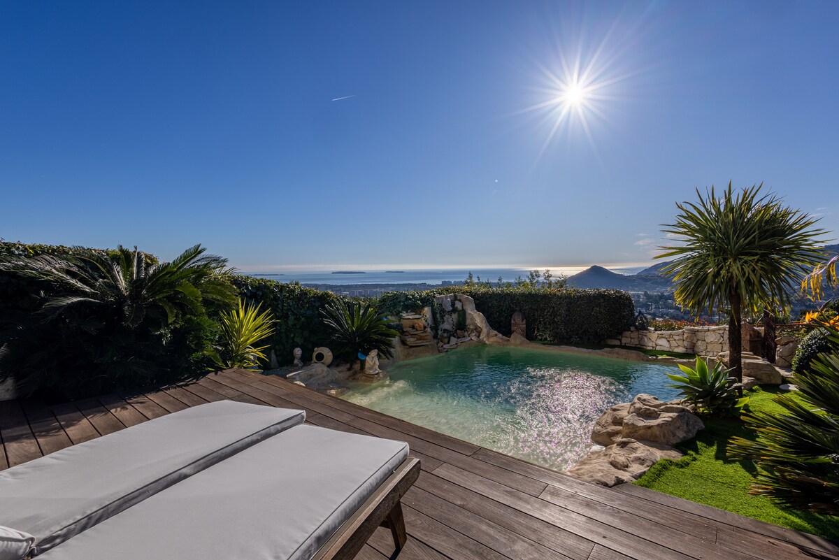 A private pool is surrounded by tropical plants and a wooden deck. The sun shines brightly above, reflecting off the water's surface. The expansive view includes the Mediterranean Sea and distant hills, creating a serene outdoor space perfect for relaxation.