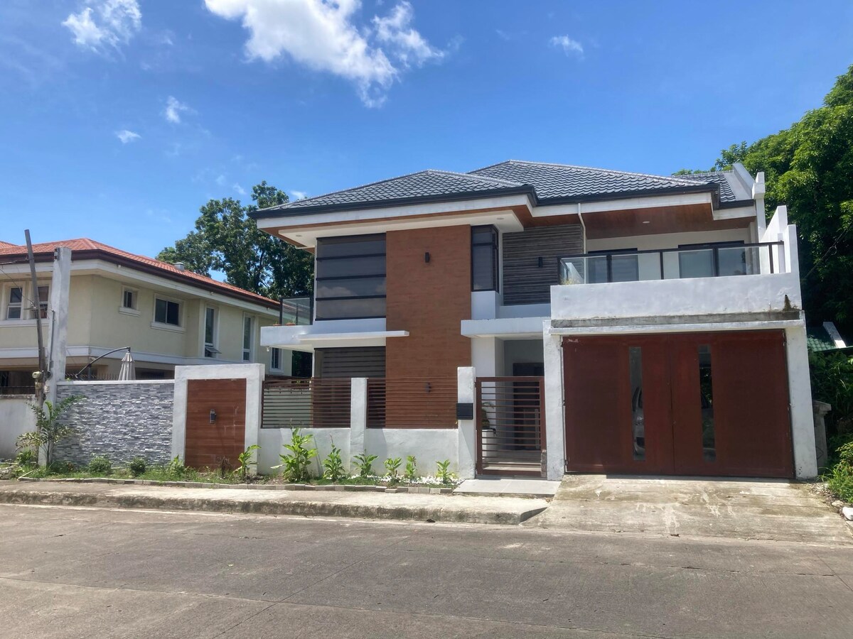 A modern two-story house features a combination of stone and wood materials on the exterior. Large windows allow natural light to fill the interior. The entrance is framed by a sleek fence and a landscaped area, with a paved driveway and surrounding greenery.