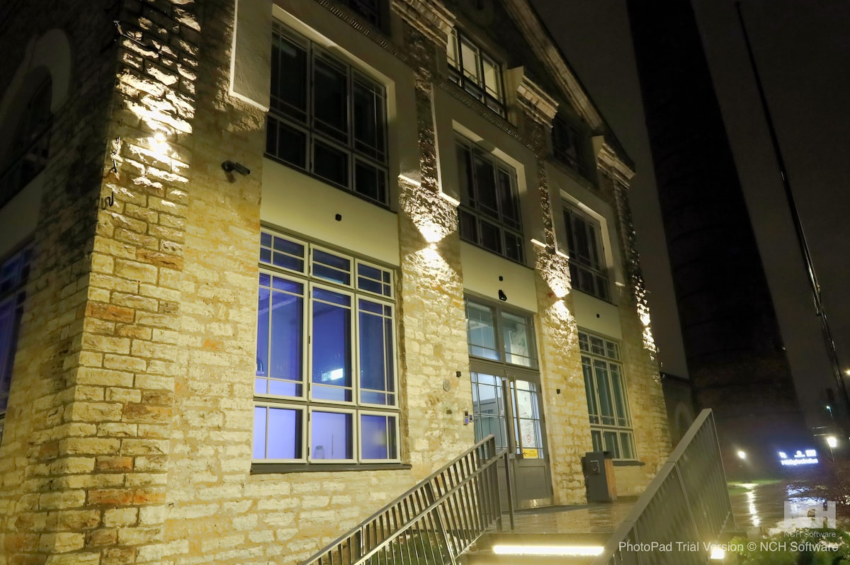 The exterior of a historical building is illuminated at night, highlighting its stone facade and large windows. A welcoming entrance is visible, featuring a set of stairs leading up to the main door. The surrounding area is well-lit, enhancing visibility.