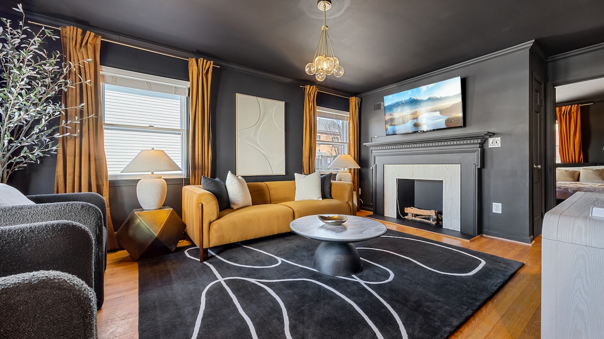 A stylish living area features a mustard-colored sofa complemented by cream and dark grey accents. The space is lit by a modern chandelier and natural light from two windows. An abstract area rug anchors the room, with a circular coffee table placed centrally. A decorative fireplace adds charm.