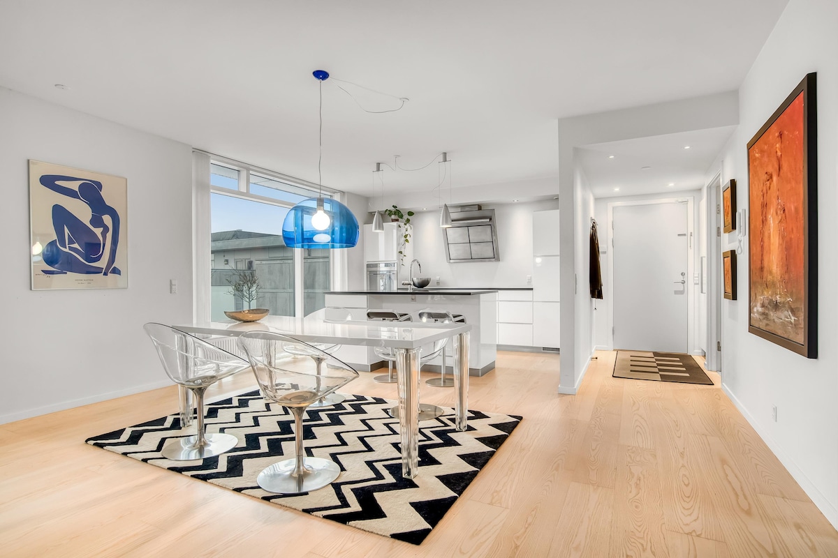 A modern kitchen area with sleek white cabinetry and a glass dining table is featured. The space is accented by transparent chairs and a bold patterned rug beneath. Natural light floods through large windows, creating a bright and airy atmosphere.