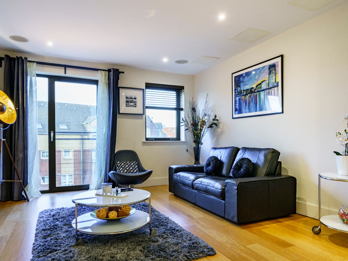 A bright living room is presented, featuring a comfortable black sofa facing a coffee table. A large window with sheer curtains allows natural light to filter in. Decorative plants and a piece of artwork add a touch of elegance to the space, complemented by a soft area rug.