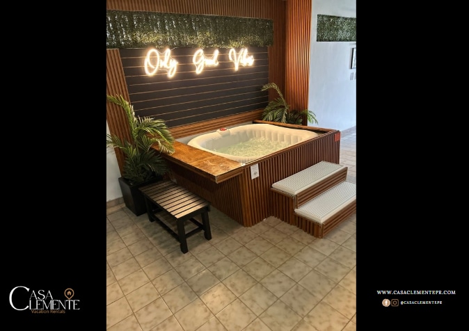 A private jacuzzi is situated in a stylish wooden alcove with modern wall decor featuring the phrase 'Only God Vibe.' Tropical plants add greenery, and a wooden bench is placed nearby. The tiled flooring complements the serene setting.