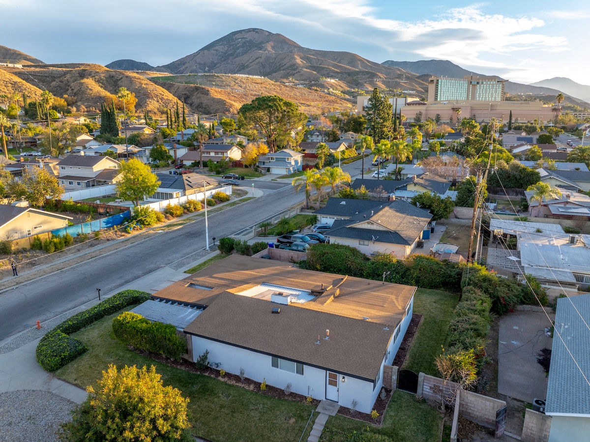 An aerial view captures a peaceful residential neighborhood, showcasing the single-story house with a beige roof. Lush greenery surrounds the property, while distant hills and mountains create a scenic backdrop. The surrounding roads and parked vehicles can also be observed.