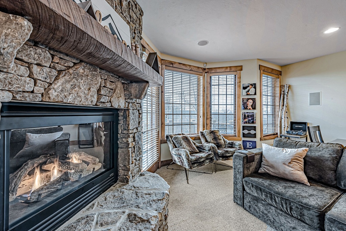 A cozy living area is highlighted by a stone fireplace, with a soft glow from the flames. Two patterned armchairs and a plush sofa provide comfortable seating, while large windows offer natural light and scenic views. The room features a warm, inviting color palette.
