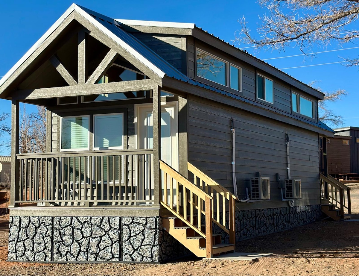 A tiny home exterior is showcased, featuring a sloped roof and a covered porch with wooden railings. Large windows provide ample natural light, while two air conditioning units are visible on the side. Stairs with handrails lead up to the entrance.