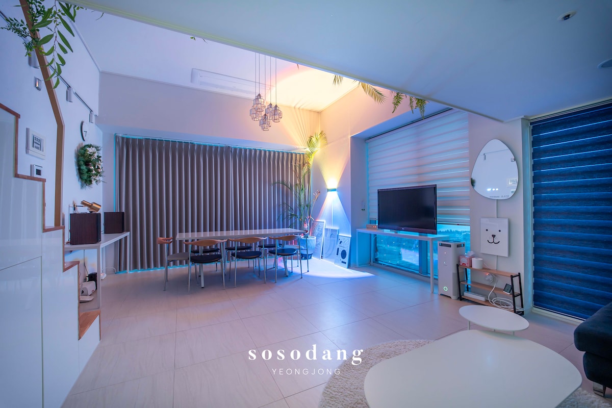 A spacious living area features a large marble dining table surrounded by eight chairs. A comfortable four-person sofa is positioned nearby, complemented by a coffee table. A smart TV is available for entertainment, along with controlled lighting and automated curtains for a pleasant atmosphere.