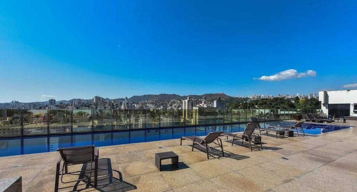 A rooftop area showcases a clearer view of the city skyline and surrounding hills. Multiple lounge chairs are positioned along the edge of a shimmering swimming pool, with a glass railing ensuring safety. The space is illuminated by natural sunlight under a clear blue sky.