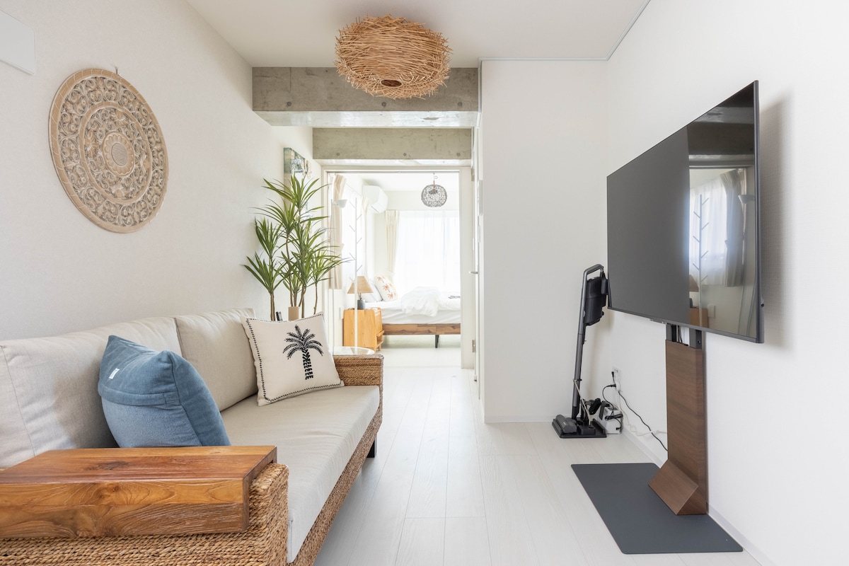 A light-filled living area features a comfortable sofa adorned with decorative cushions. A large flat-screen TV stands on a sleek wooden stand. A potted plant adds a touch of greenery, while a textured ceiling light contributes to the room's design.