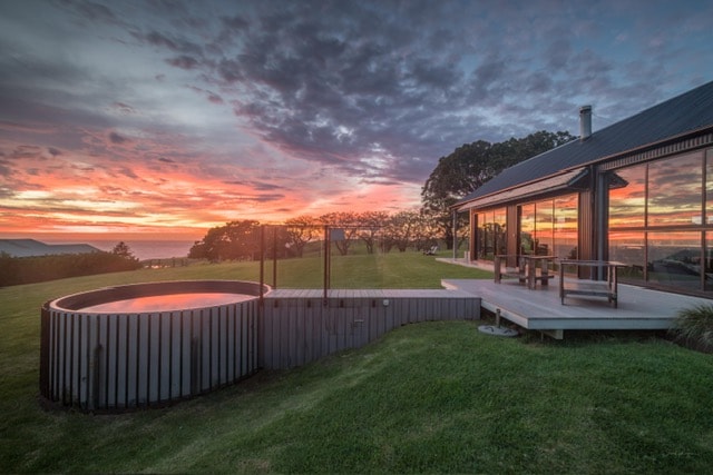 A modern retreat with a spacious deck overlooks rolling green lawns leading to a private plunge pool. The setting sun casts vibrant hues across the sky, while large glass windows reflect the natural beauty and enhance indoor-outdoor connection.