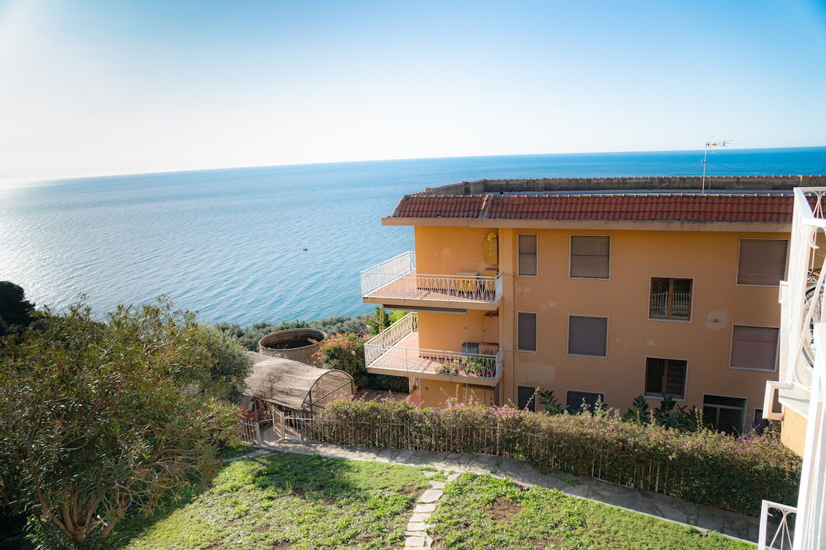 The image showcases an expansive view of an apartment building situated on a hillside, overlooking the sea. The structure features multiple floors and balconies, surrounded by greenery and a well-maintained garden area. The tranquil blue waters stretch to the horizon, illuminated by natural light.