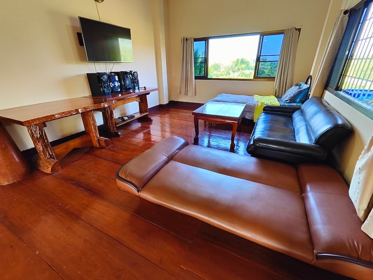 A living area features a dark leather sofa and a wooden coffee table. Large windows provide natural light and views of the surrounding landscape. A wooden console accommodates a flat-screen TV, enhancing the space's comfort and functionality.