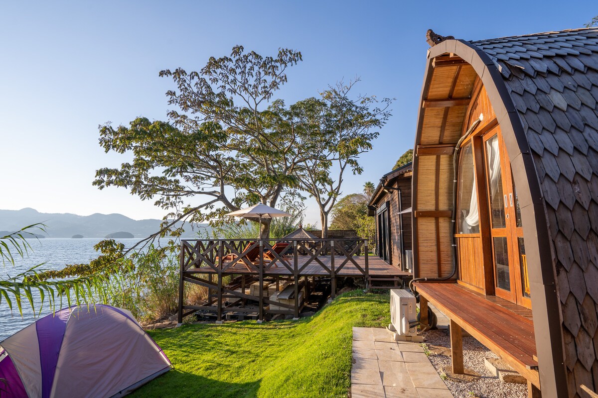An inviting glamping site features a wooden structure with large windows, overlooking a scenic waterfront. A spacious deck is visible, furnished with seating options, while vibrant green grass surrounds the area. Nearby, a colorful tent adds to the outdoor atmosphere under a clear blue sky.
