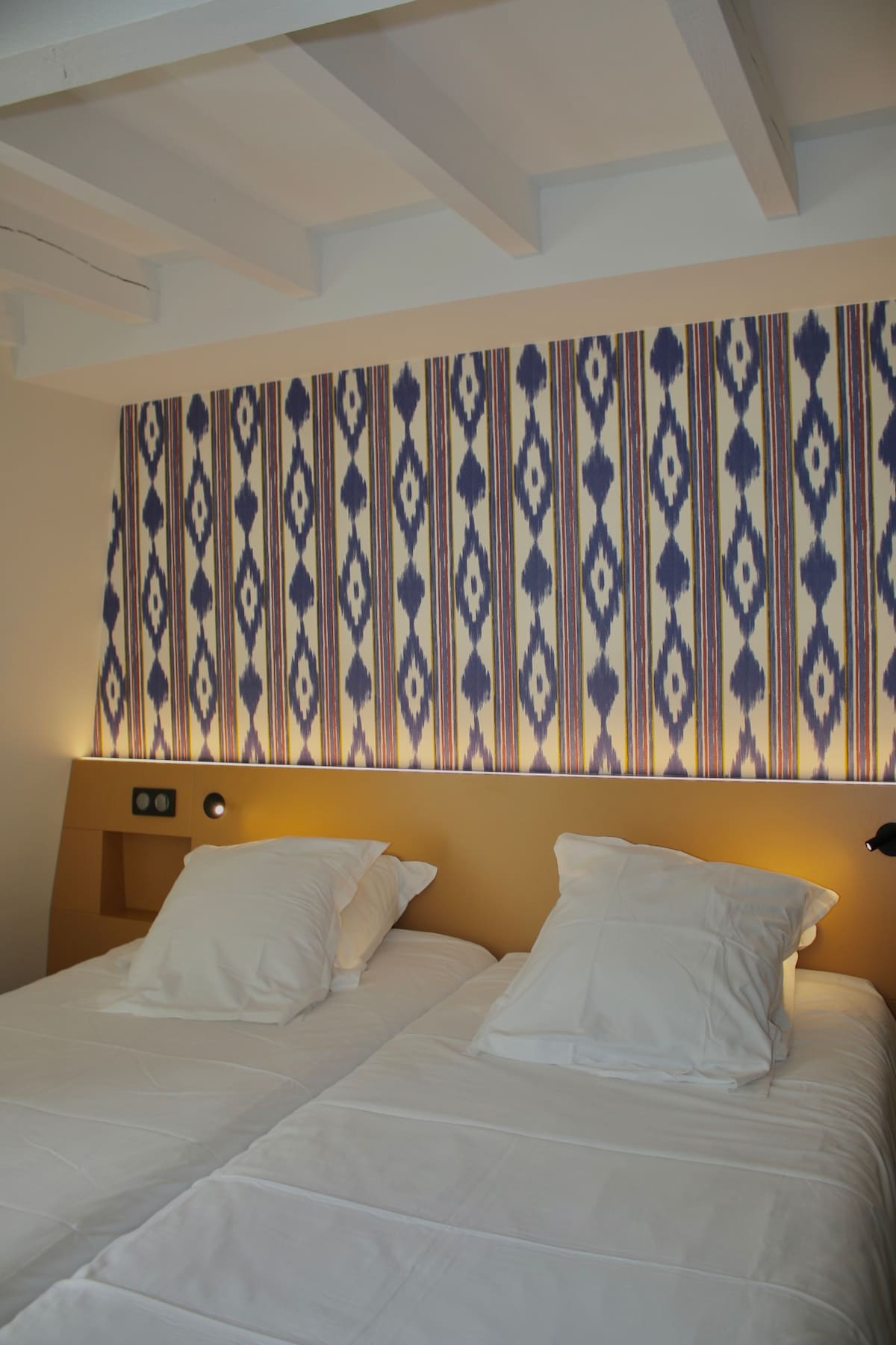 The room features two spacious twin beds dressed with crisp white linens and plush pillows. A patterned accent wall in blue and cream adds visual interest. Soft lighting is provided by wall-mounted sconces beside each bed.