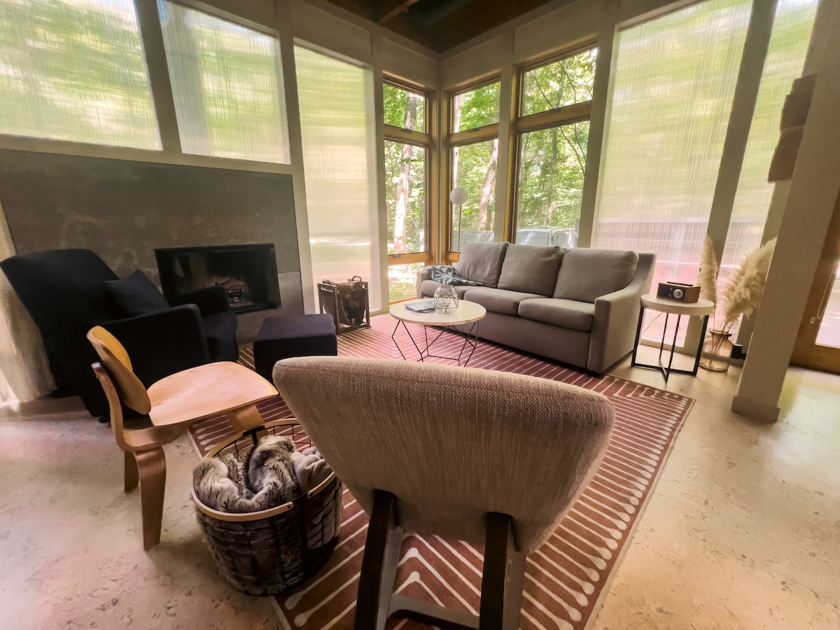 A bright living area features large windows allowing natural light to fill the space. A comfortable gray sofa is positioned near a modern fireplace, accompanied by a couple of armchairs. A coffee table sits atop a patterned rug, inviting relaxation.